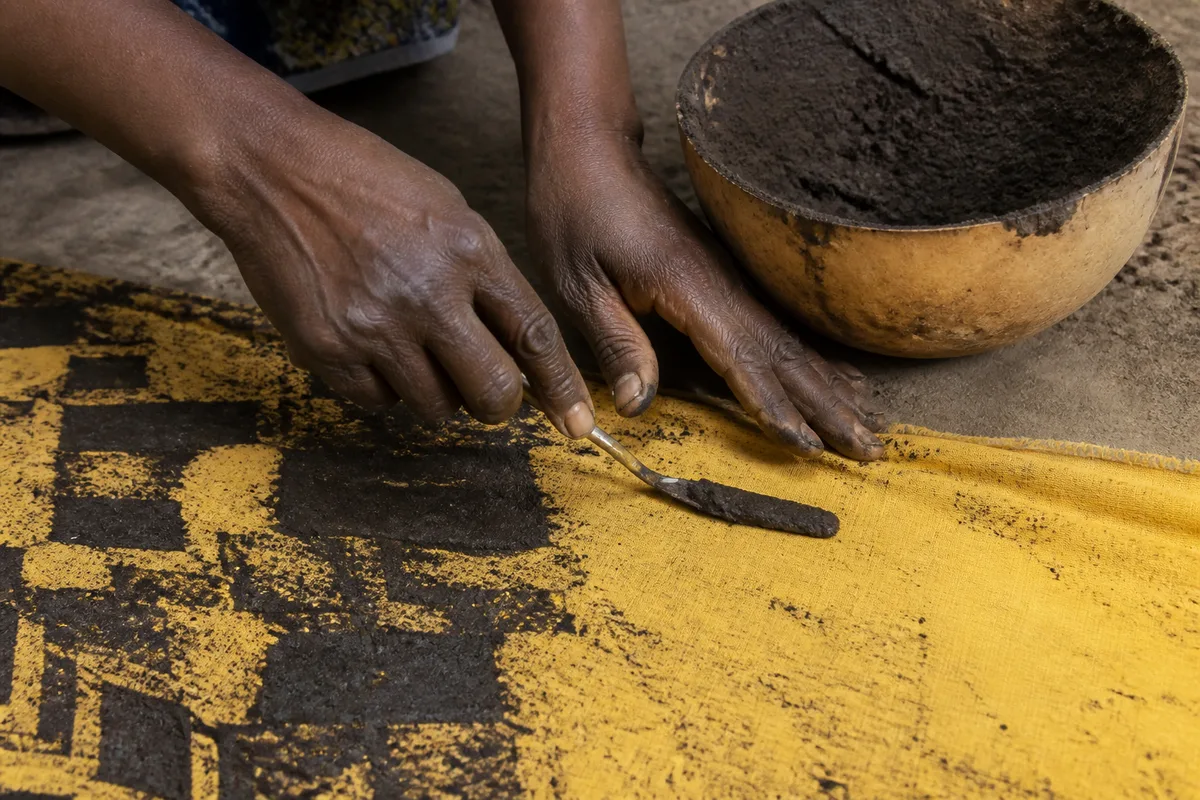 Mains d&rsquo;artisane appliquant de la boue fermentée sur un coton jaune avec une fine spatule métallique, atelier en plein air