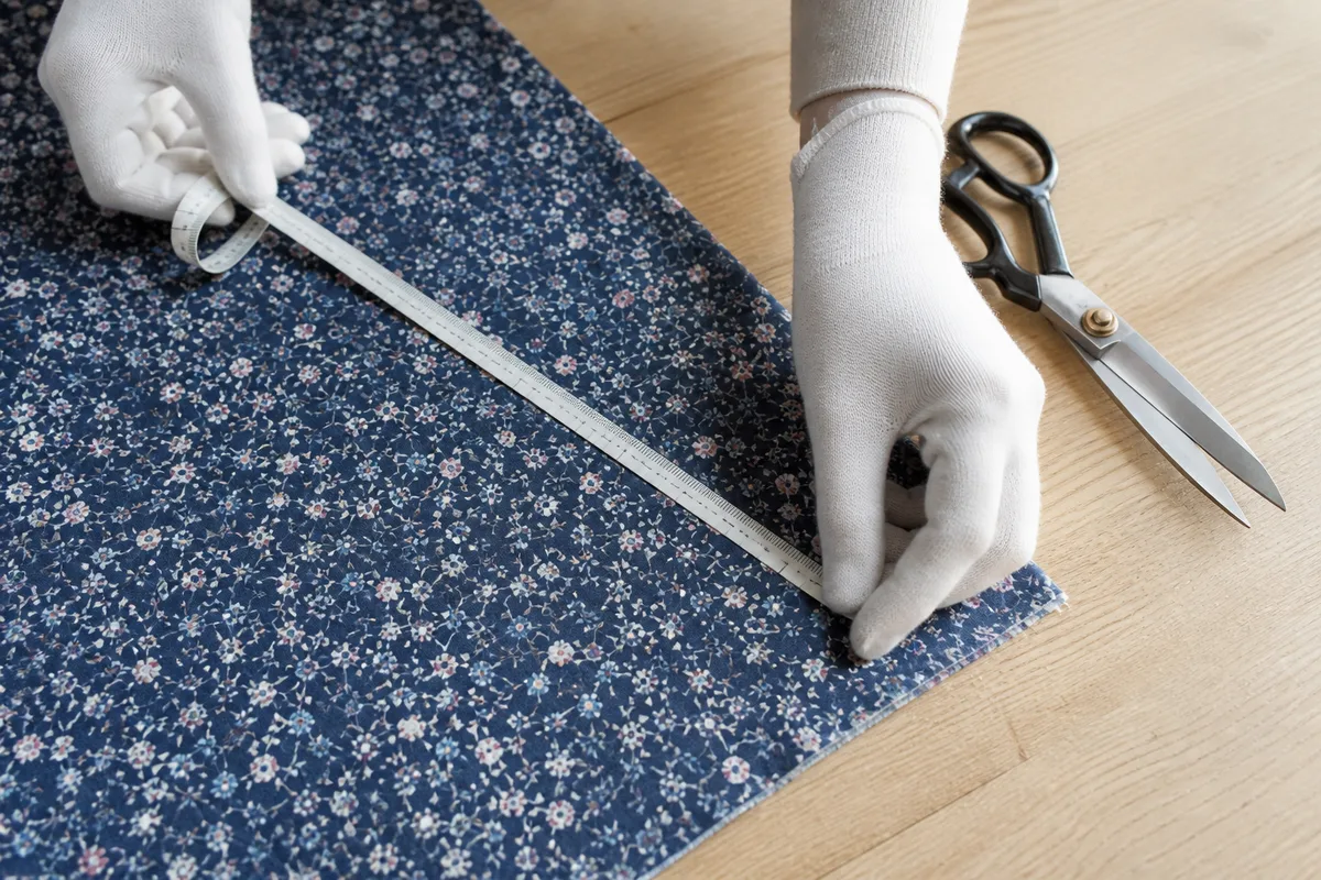 Employée gantée mesurant un métrage de tissu sur une table de coupe en bois clair, mètre ruban déroulé